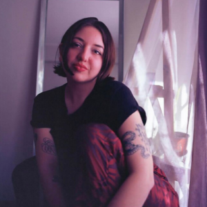 Photo of a woman with short brown hair wearing a black top and red pants, looking off camera with legs crossed