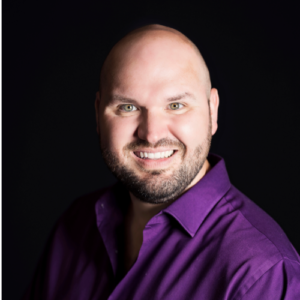 Photo of man wearing purple button up shirt, smiling directly at camera